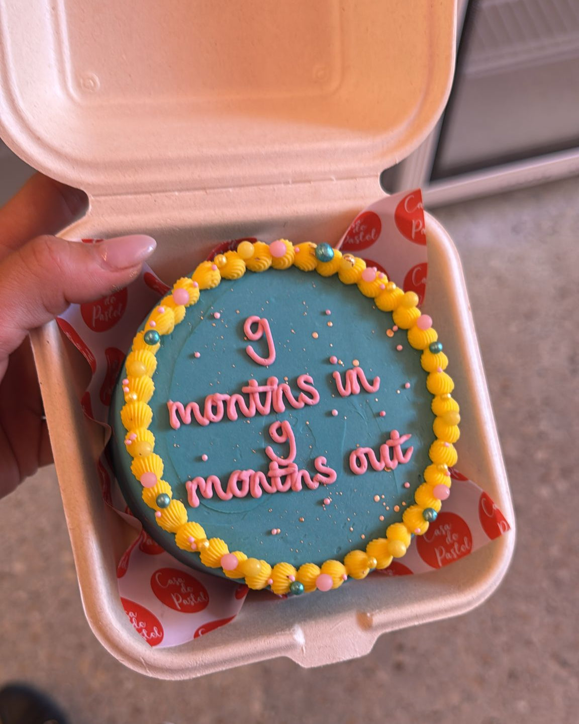 Small round cake with '9 months in 9 months out' text on a blue background, held in a takeout container.