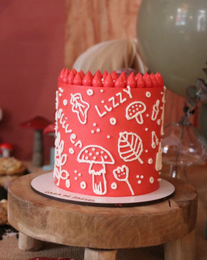 Red cake with white designs on a wooden stand, surrounded by decorative mushrooms.